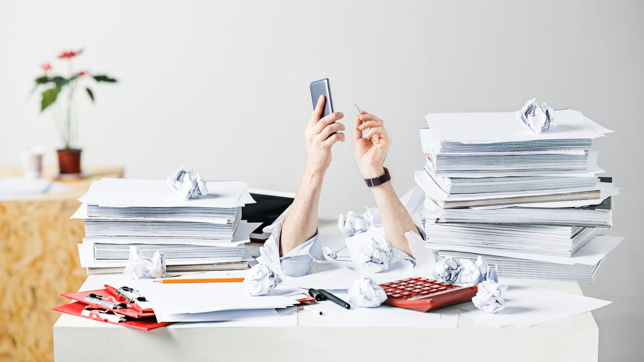 A woman is sitting at a desk with a pile of papers and a cell phone.
