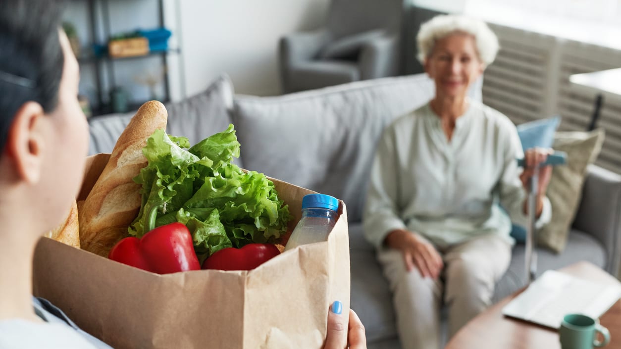 A woman is holding a paper bag full of groceries in front of a couch.
