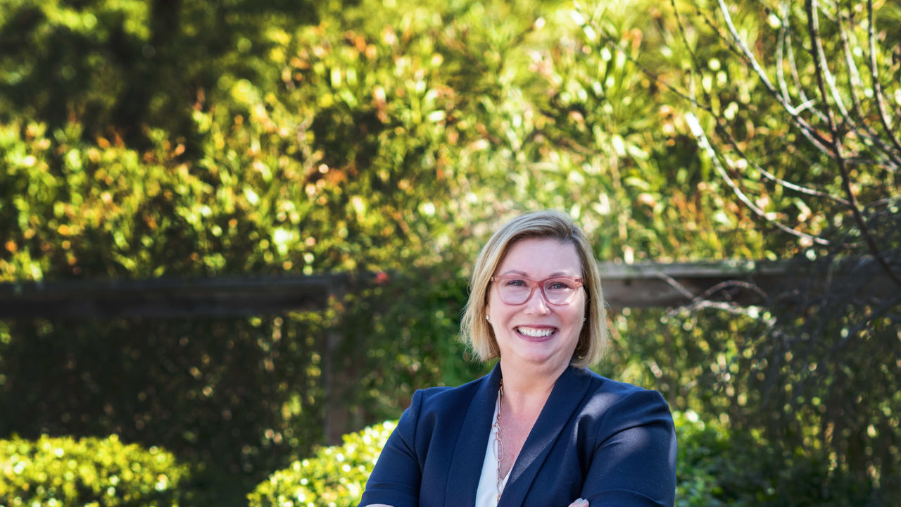 A woman in jeans and a blazer standing in front of bushes.