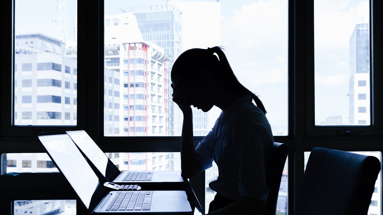 A woman sitting at a desk with two laptops in front of a window.