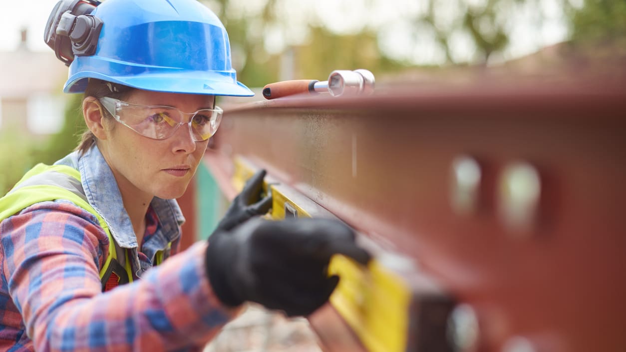 A woman wearing a hard hat and safety glasses is working on a metal beam.