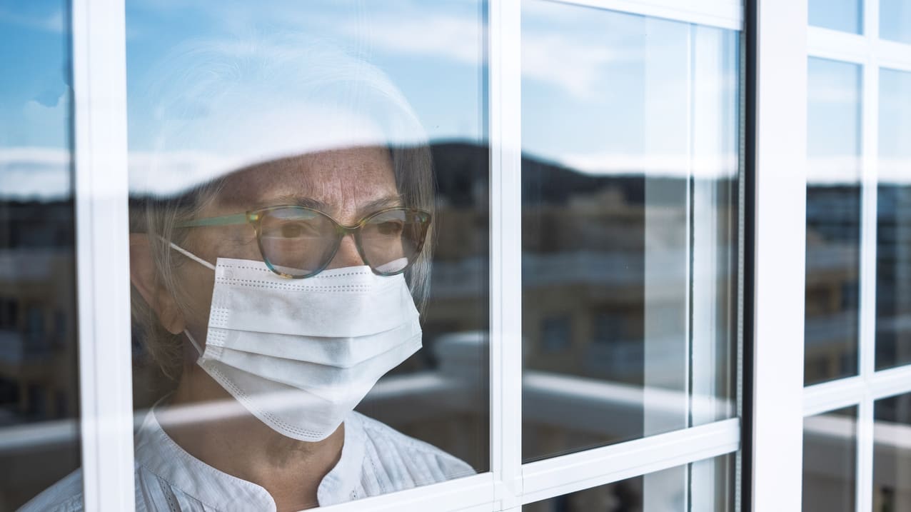 A woman wearing a surgical mask looking out of a window.