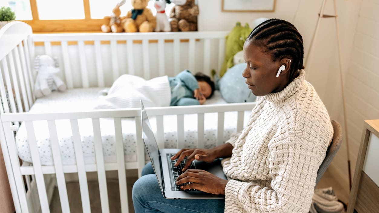 A woman using a laptop in front of a baby's crib.