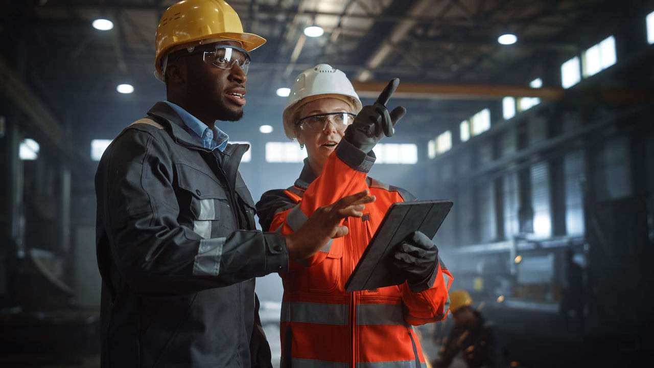 Two workers in hard hats looking at a tablet in a factory.