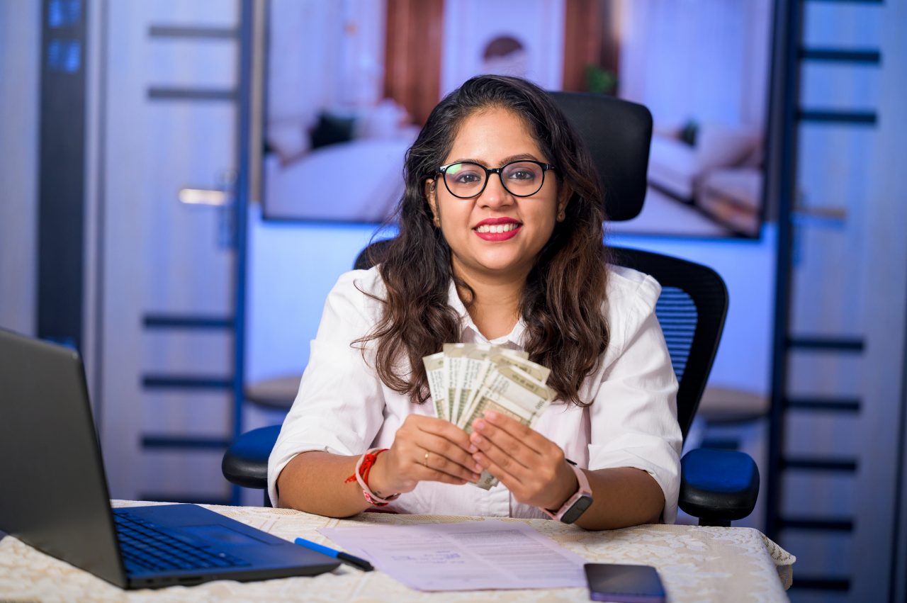 Cheerful young woman holding paper currency while sitting on office chair