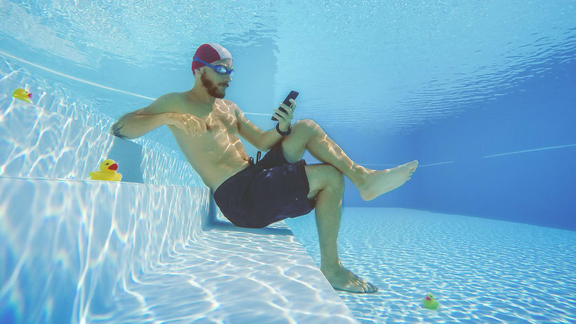 A swimmer holding a phone underwater with rubber ducks nearby.
