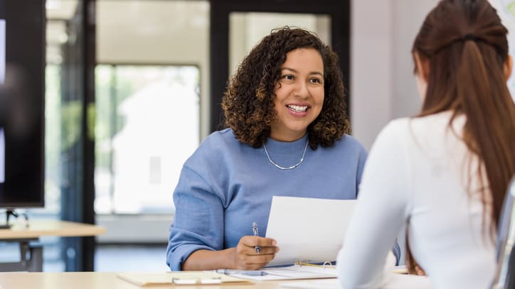 Two women sitting at a desk talking to each other.