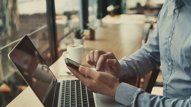 A man is using his phone while sitting at a table with a laptop.