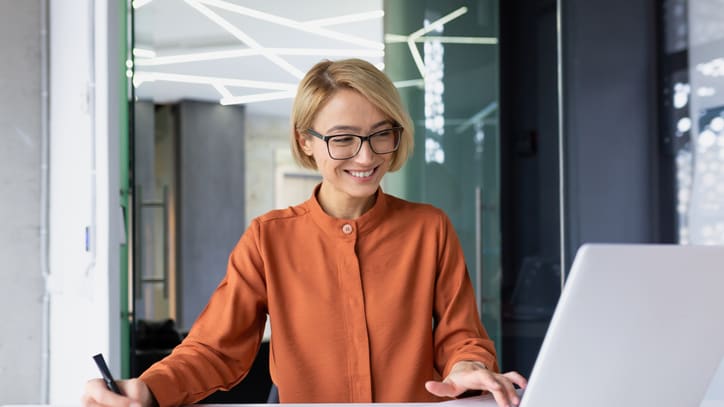 A woman in glasses is sitting at a desk with a laptop.