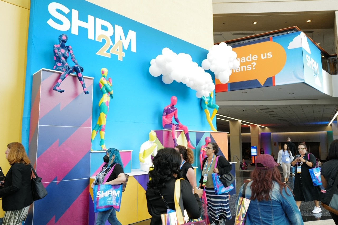Attendees at SHRM24 walk past a colorful sign painted with a blue sky and balloon clouds