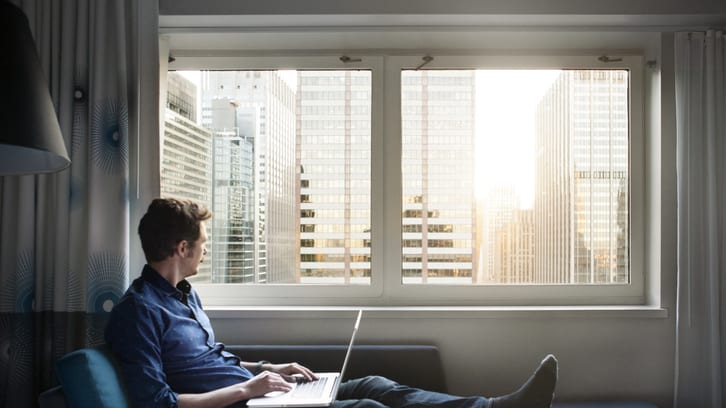 A man sitting on a bed with a laptop in front of a window.