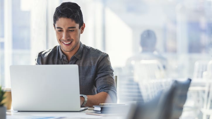 A man is smiling while working on a laptop in an office.