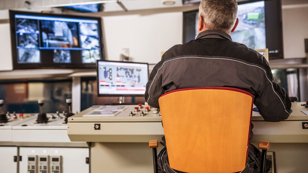 A man sitting at a control room with several monitors.