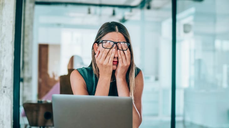 A woman covering her eyes while working on a laptop.