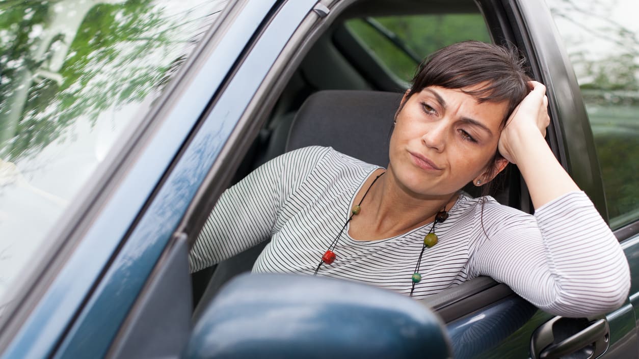 A woman sitting in a car with her head resting on her hand leaning out the window.