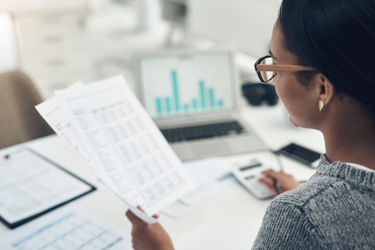 A woman wearing glasses looks at a paper budget spreadsheet and a computer with a bar graph on the screen. 