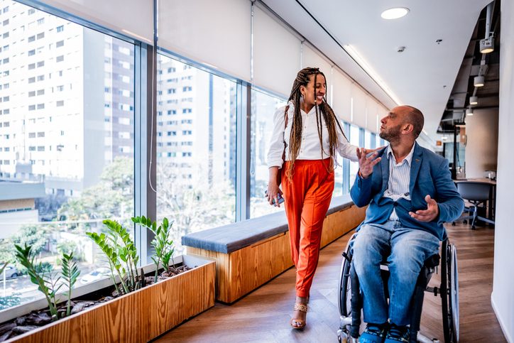 A man in a wheelchair talks to a female colleague as they go through a hallway at the office.