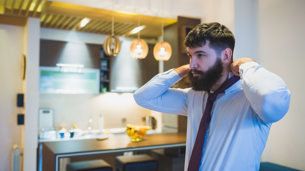 A man with a beard is adjusting his tie in a living room.