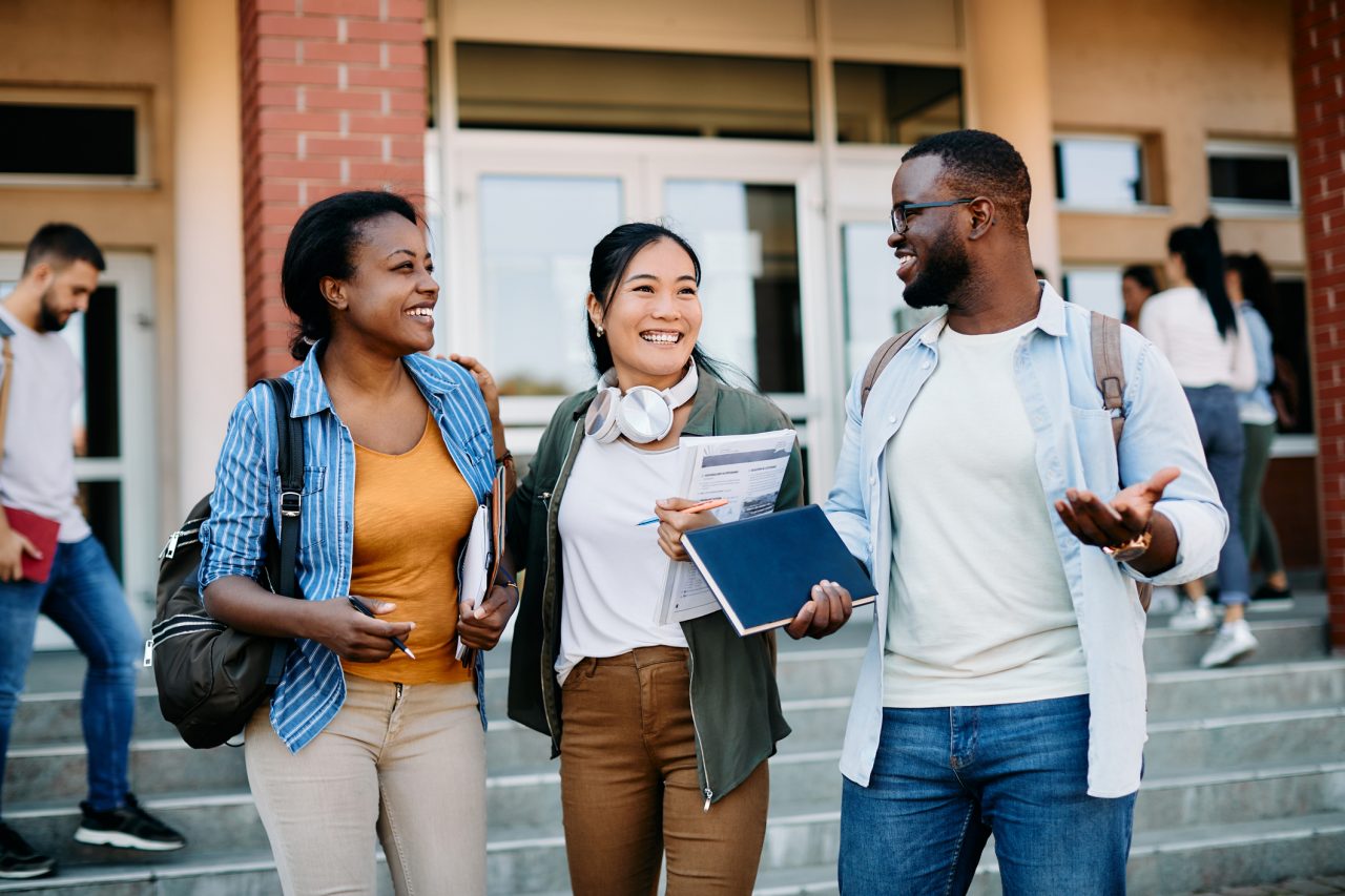 Three college students smiling and talking while walking down steps outside a school building.