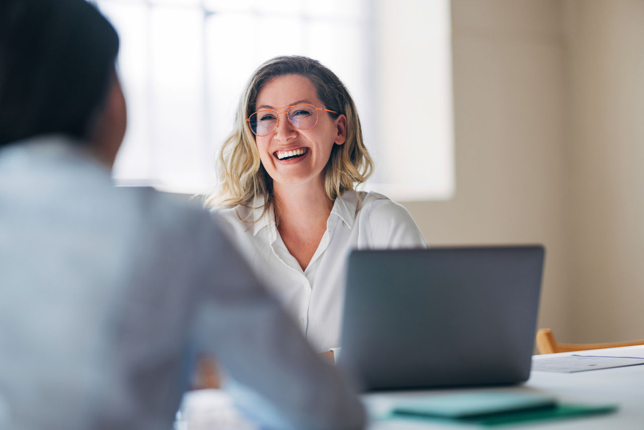 A cheerful woman with glasses is sitting at a desk, engaged in a friendly business meeting. The bright office setting creates a positive and collaborative atmosphere.