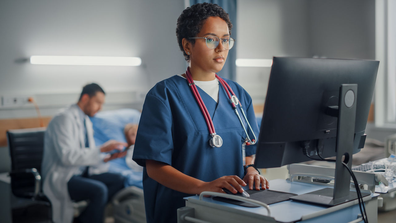 A nurse at a computer with a doctor and patient in the background.