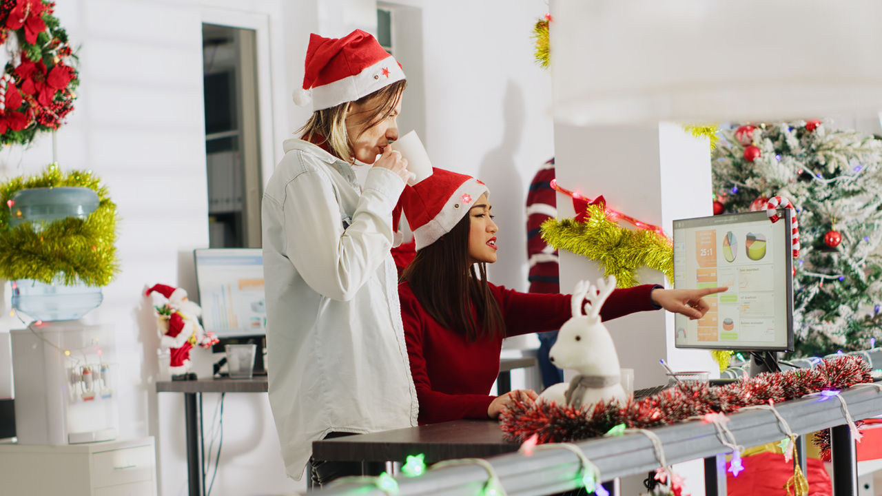 Employee wearing Santa Claus hat working in Christmas ornate office and chatting with coworker. Worker looking over business revenue numbers at computer desk in xmas adorn workplace, handheld camera