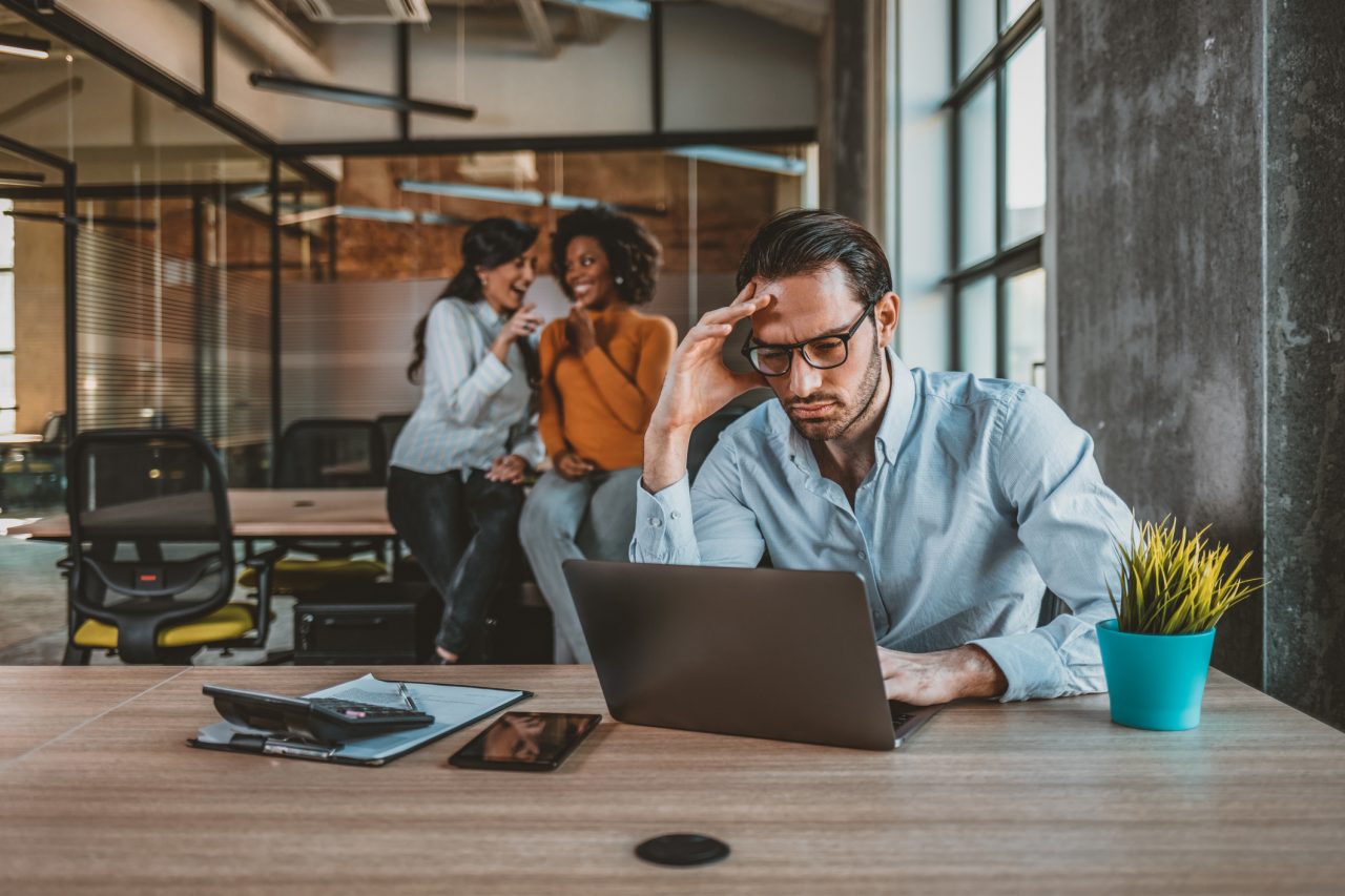 Man sits at computer while female coworkers gossip behind him