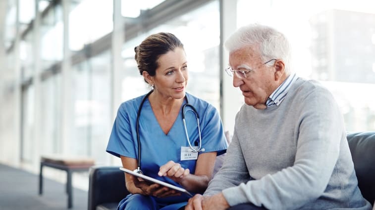 A nurse is talking to an older man on a couch.