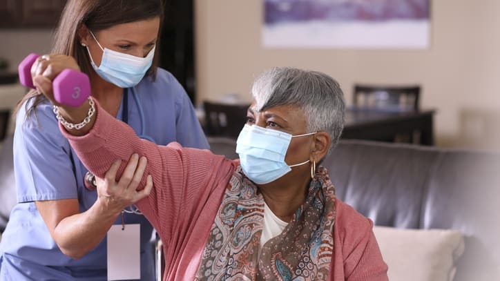 A nurse wearing a face mask is helping an elderly woman lift weights.