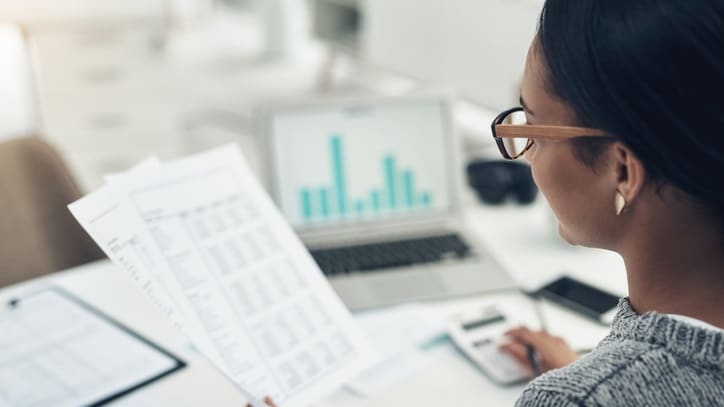 A woman in glasses is looking at a piece of paper at her desk.