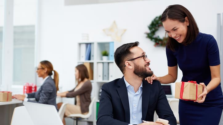 A woman is holding a gift in front of a man in an office.