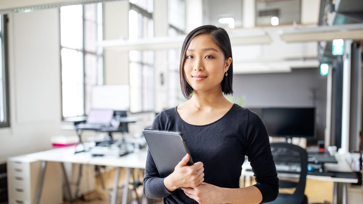 A young woman standing in an office holding a tablet computer.