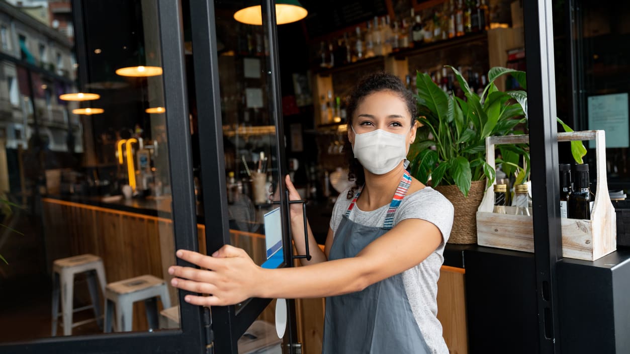 A woman wearing a face mask is opening the door of a restaurant.