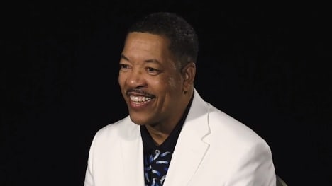 A man in a white suit is smiling in front of a black background.