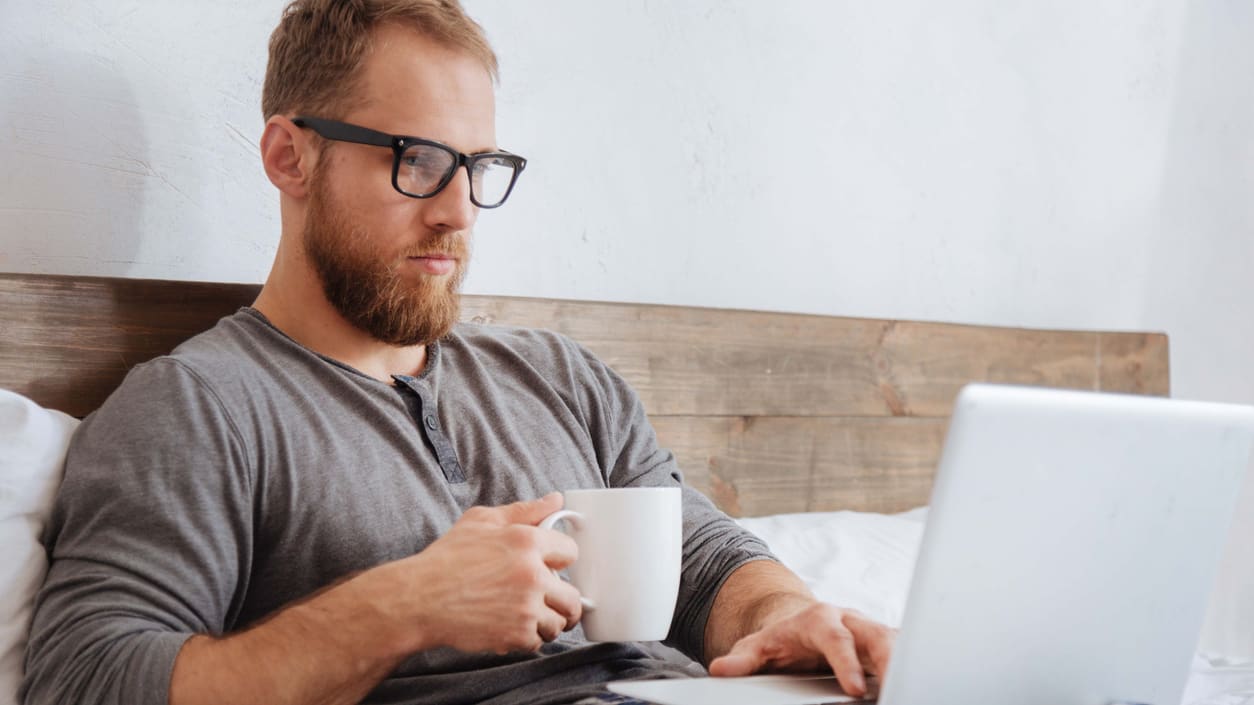 A man using a laptop in bed with a cup of coffee.