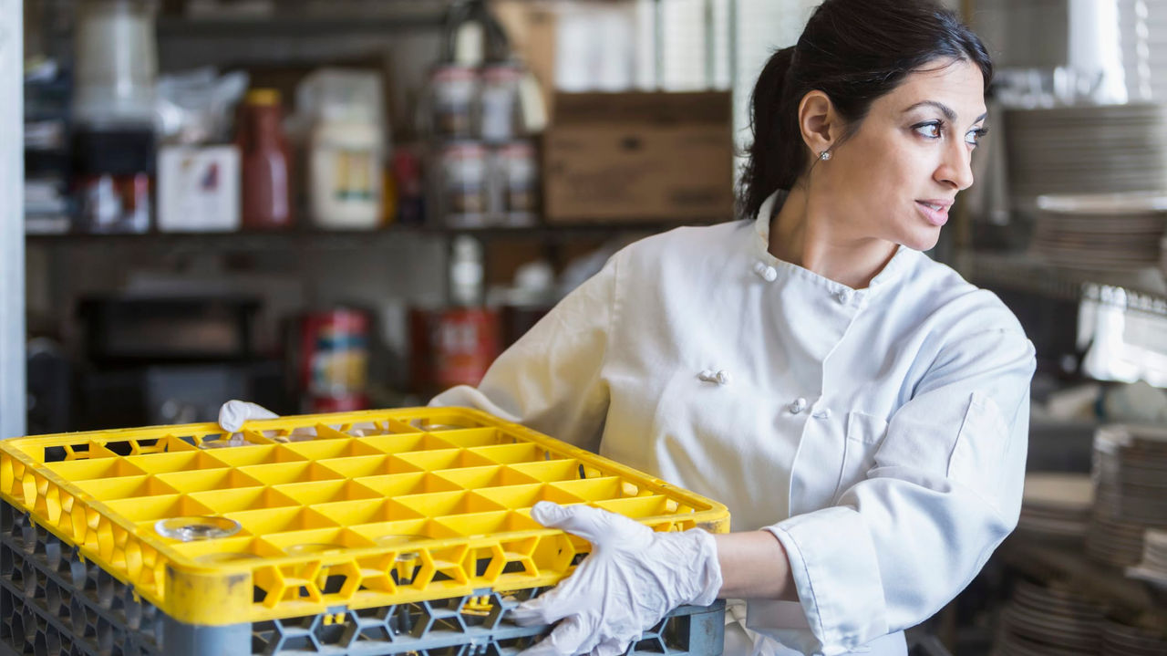 A female chef holding a yellow crate in a kitchen.