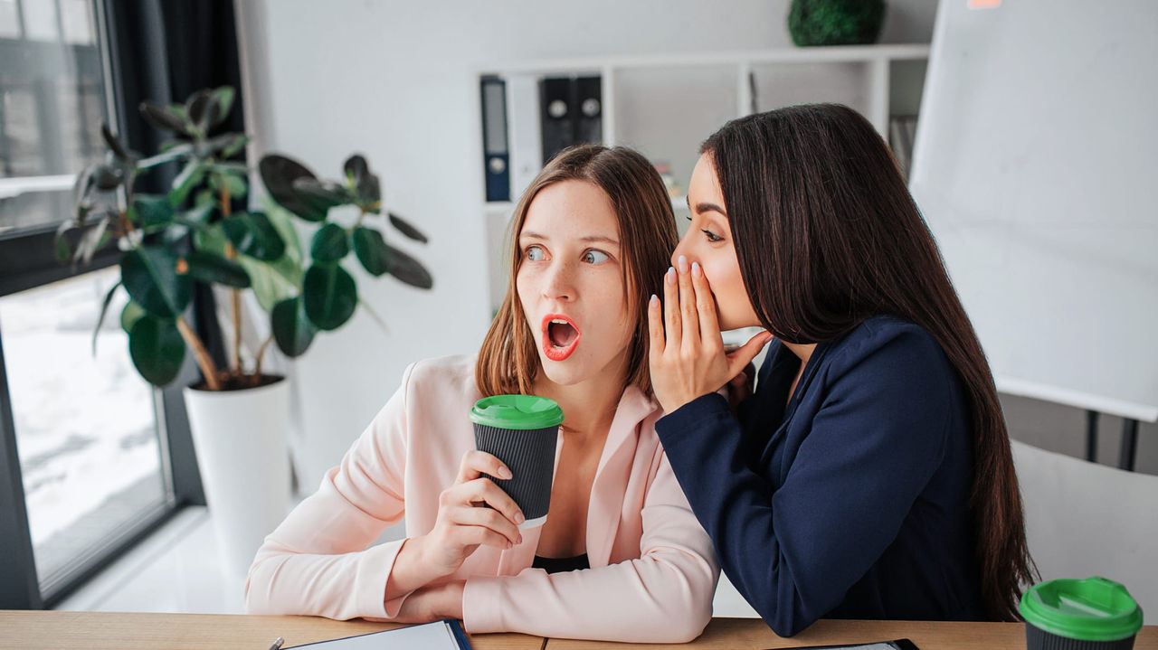 Two women in an office with a cup of coffee and a mouth open.