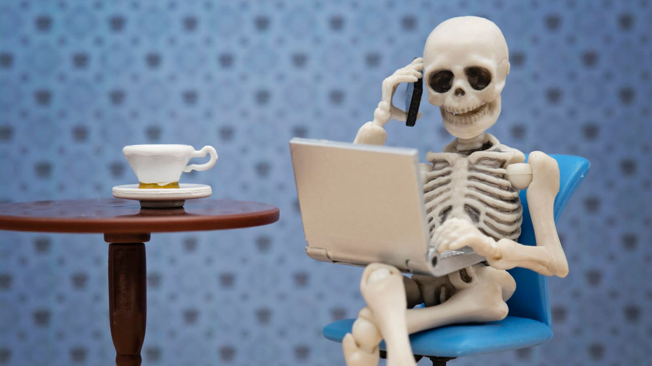 A skeleton sitting in a chair with a laptop and a cup of coffee.