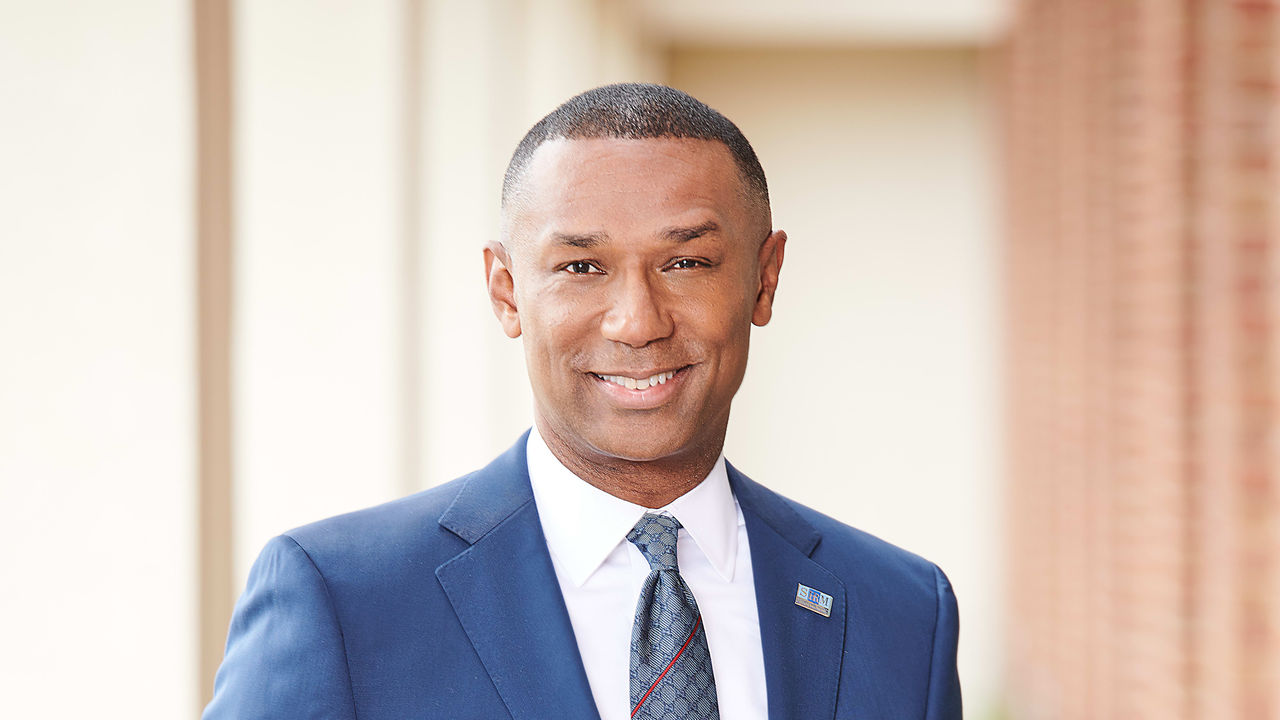 A man in a blue suit smiling in front of a brick wall.