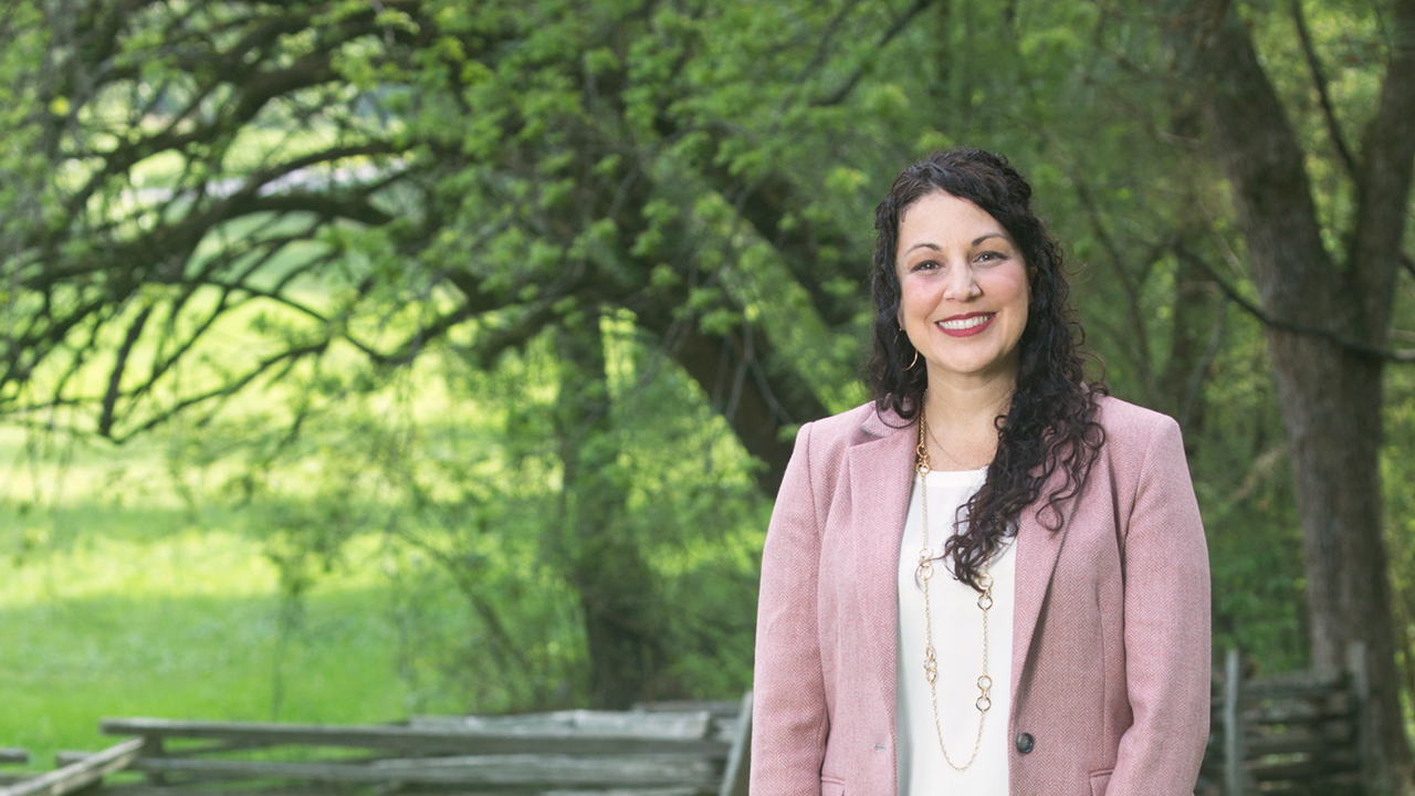 A woman in a pink blazer standing in front of a wooded area.