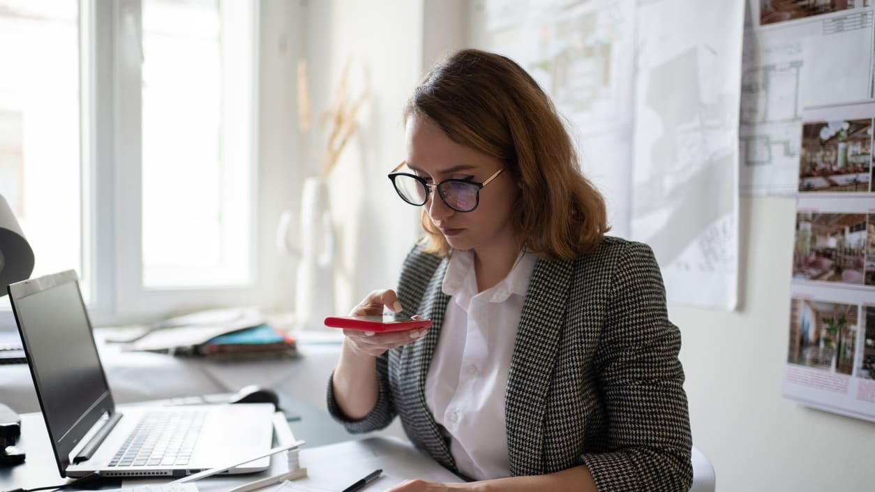 A woman in glasses is looking at her phone while sitting at a desk.