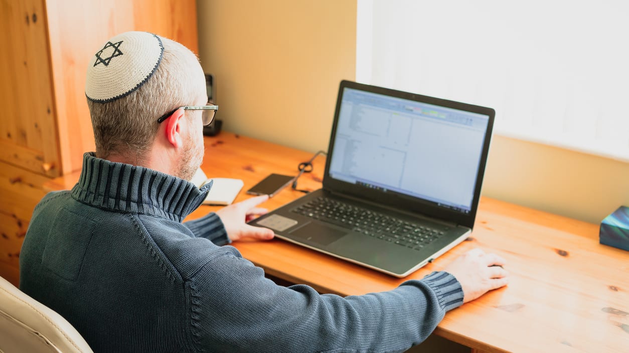 A man wearing a jewish hat is using a laptop at home.