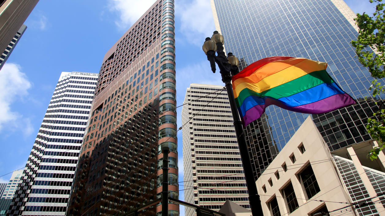 A rainbow flag is flying in front of tall buildings.