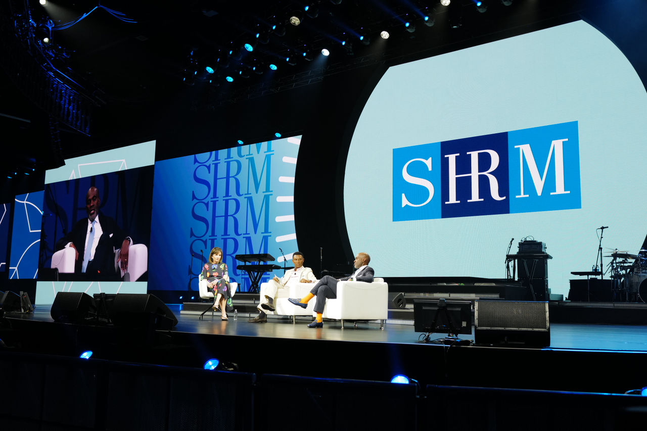 SHRM President & CEO Johnny C. Taylor, Jr.-SHRM-SCP, and SHRM board members Betty Thompson, SHRM-SCP, and Sean N. Woodroffe, SHRM-SCP are seated on stage at SHRM26, in large white chairs. They're seated in front of a large screen with the SHRM logo on it.