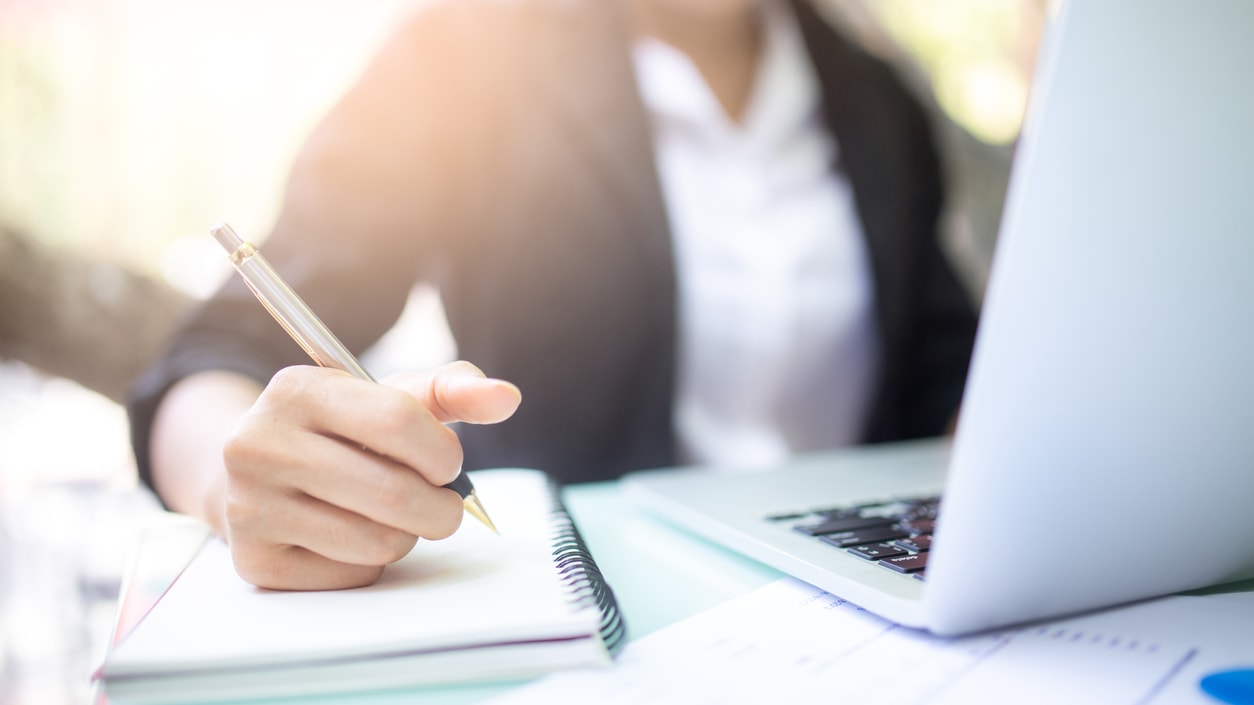 A woman is writing on a notebook while sitting at a table with a laptop.