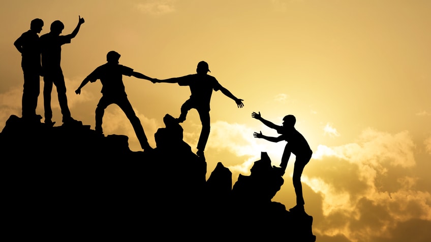 Silhouettes of people reaching for each other on top of a mountain.