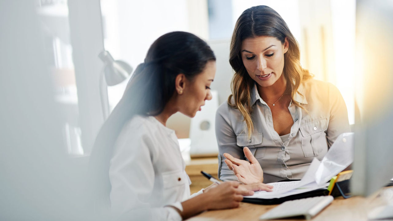 Two women sitting at a desk talking to each other.