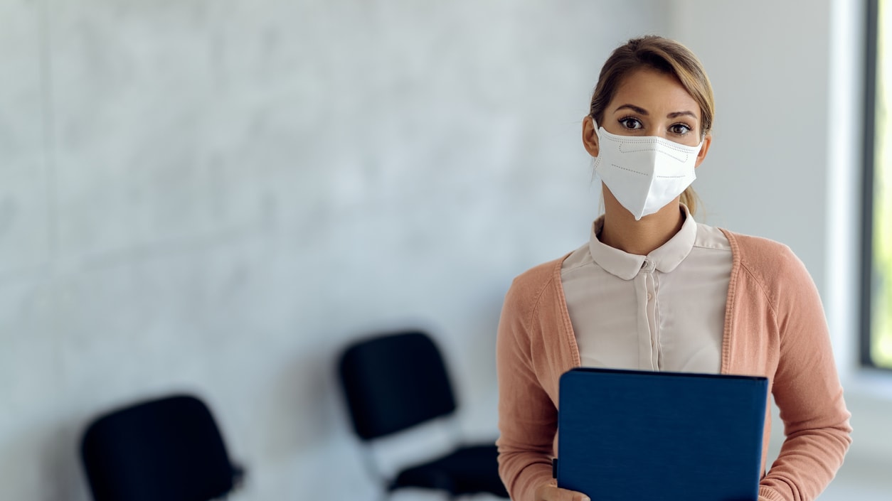 A woman wearing a surgical mask holding a folder in an office.