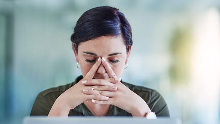 A woman covering her face with her hands while working on a laptop.