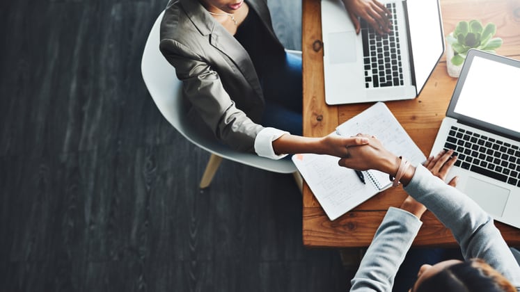 Two business people shaking hands at a table with laptops.